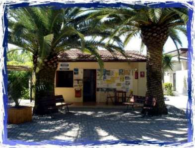 Un piccolo edificio tra palme con panchine davanti e un tabellone informativo.