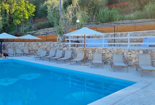 Piscina con lettini sotto ombrelloni bianchi, circondata da alberi.