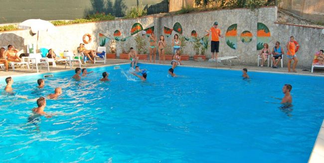 Persone giocano in piscina, con spettatori seduti sui lettini.