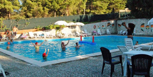 Bambini e adulti giocano in piscina, ombrelloni e sedie a bordo.