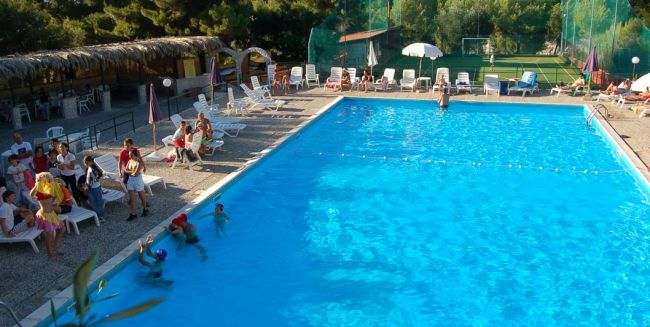 Piscina con lettini e persone sedute, immersa in un ambiente verde.