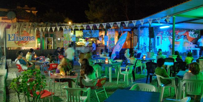 Bar all'aperto di notte con tavolini e sedie, decorato con luci colorate.