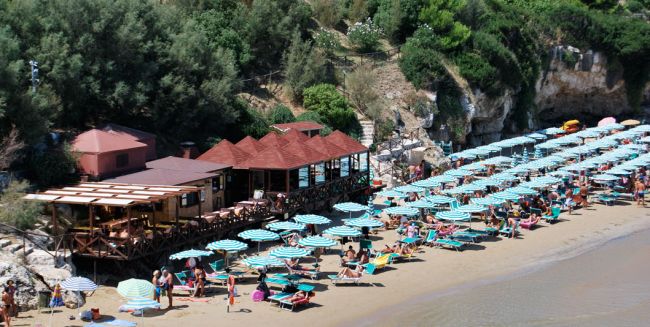 Un tratto di spiaggia con ombrelloni a righe e un ristorante sul lato, vicino a una scogliera verde.