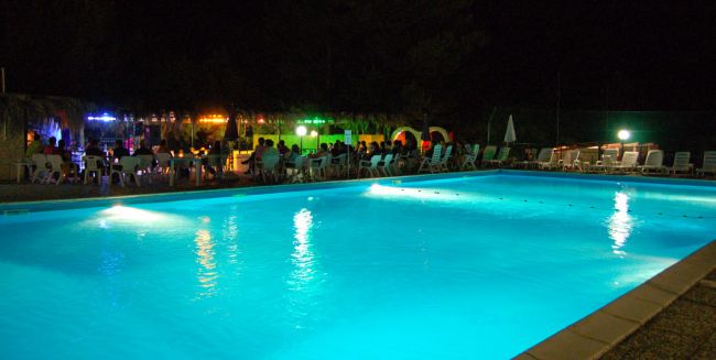 Piscina illuminata di notte con persone sedute a tavolini vicini.