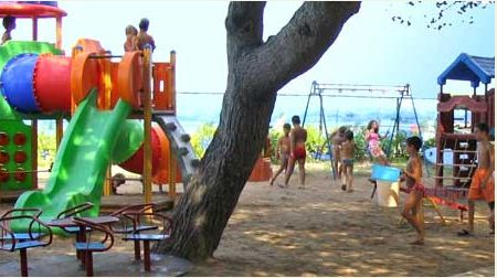 Parco giochi con scivolo colorato e bambini che giocano sotto un albero.