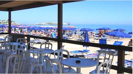 Tavolini e sedie in spiaggia con molte persone sotto ombrelloni blu e il mare sullo sfondo.