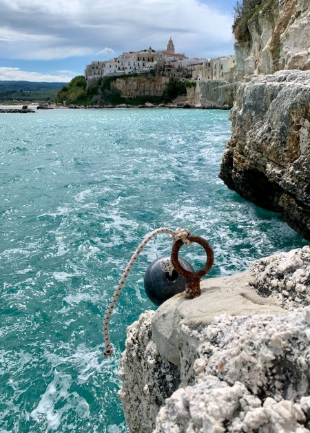 Vista di un paese su una scogliera, mare turchese e un anello d'ormeggio in primo piano.