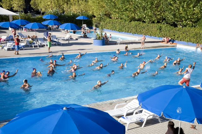 Persone fanno acquagym in piscina con ombrelloni blu.
