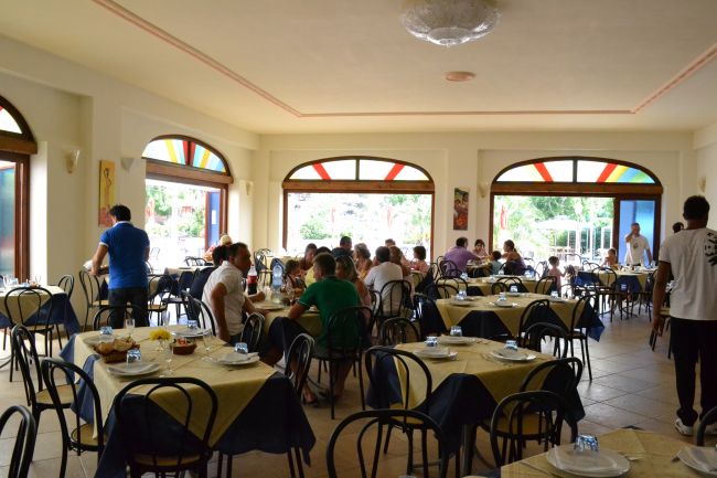 Sala da pranzo con tavoli apparecchiati, persone sedute.