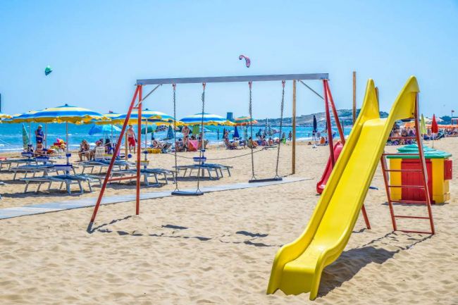 Parco giochi su una spiaggia con mare e ombrelloni.