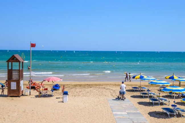 Spiaggia con passerella e torrette, ombrelloni blu e gialli.
